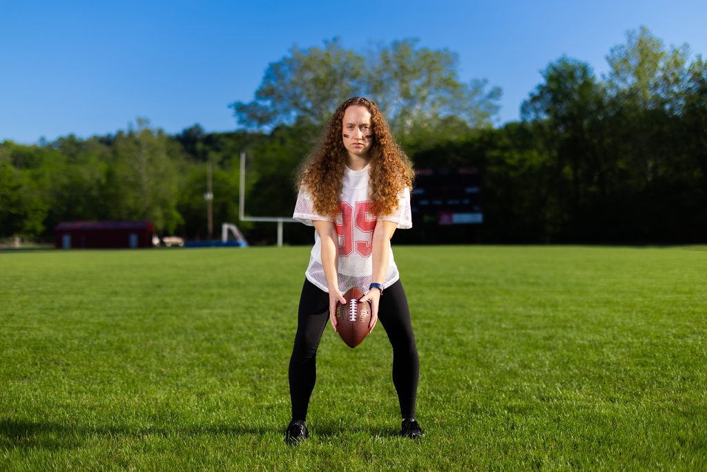 woman in football jersey holding a football in the hike position while standing on a football field