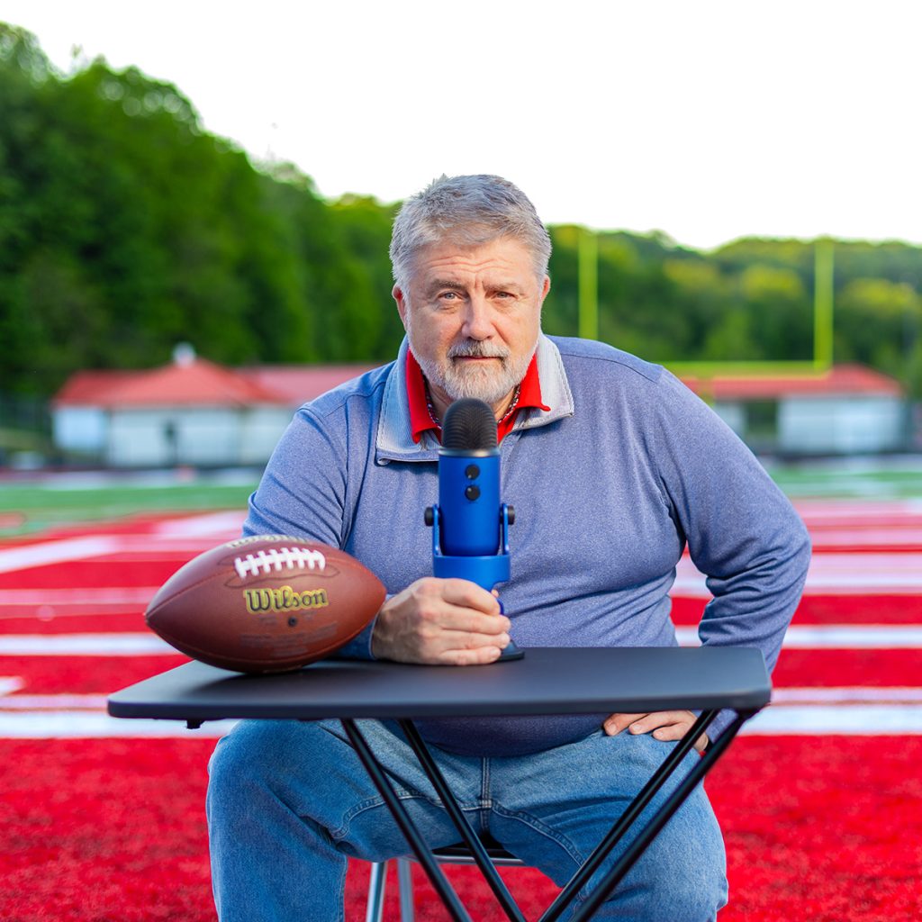 a man sitting at a small desk on a football field with a podcast microphone and football